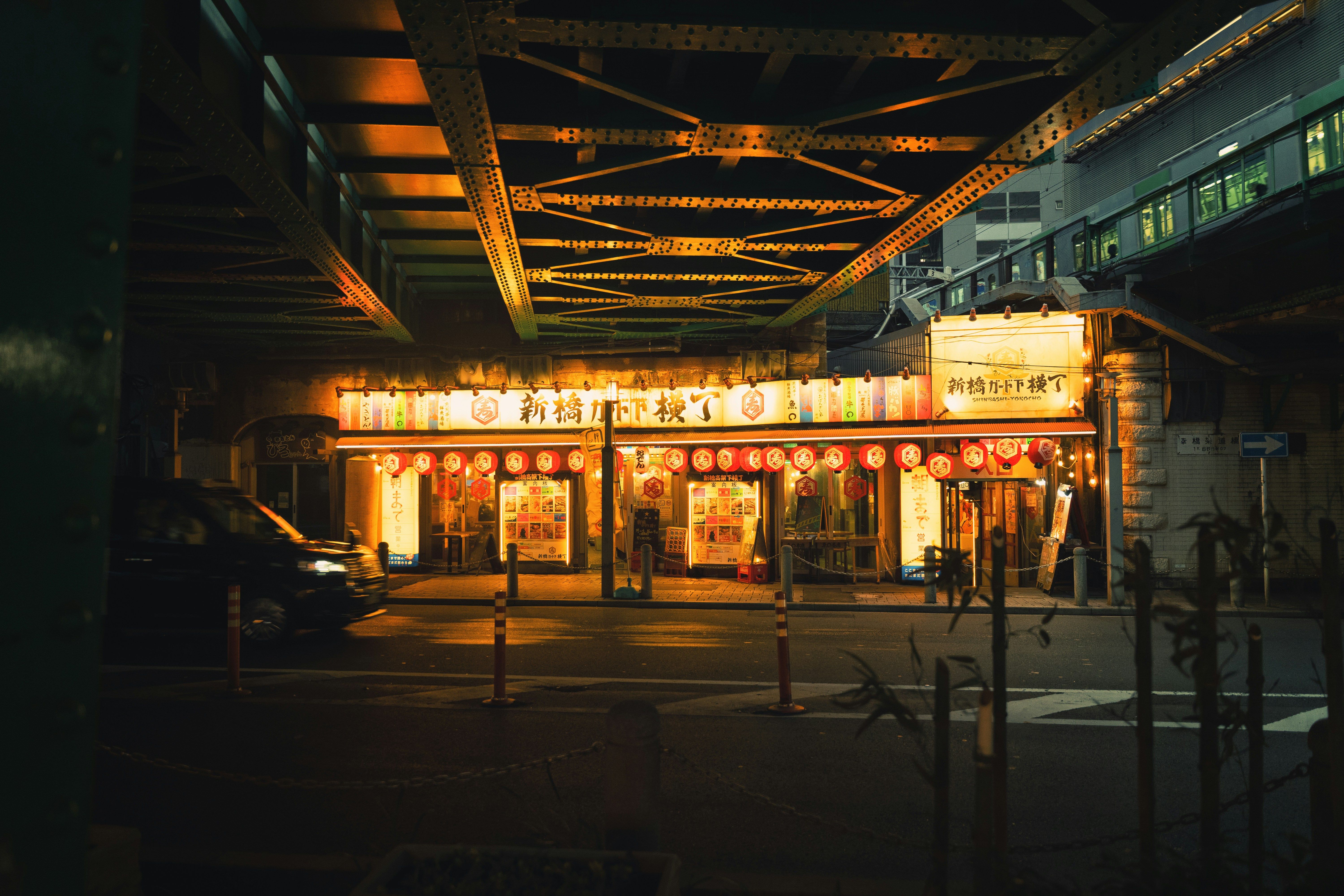 A Tokyo street in the early evening dusk. The shot is from under a steel bridge where there is a store, lit up, which in turn, lights up the surrounding area.